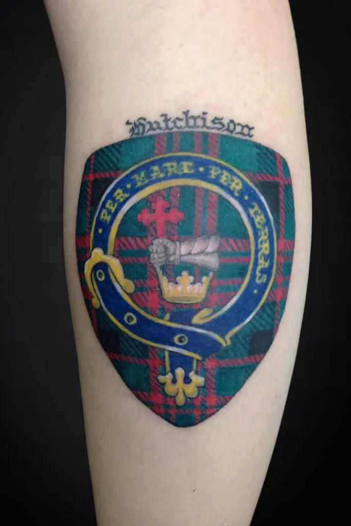 Tattoo of a heraldic shield on a plaid background, featuring a gold crown, blue ribbon, red cross, and a surrounding yellow ring, with a scripted word above the shield.