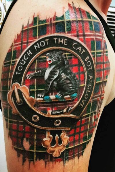 Tattoo on upper arm: a circular emblem with a rat in a leather jacket inside, set against a red-green plaid background, with text around the edge.