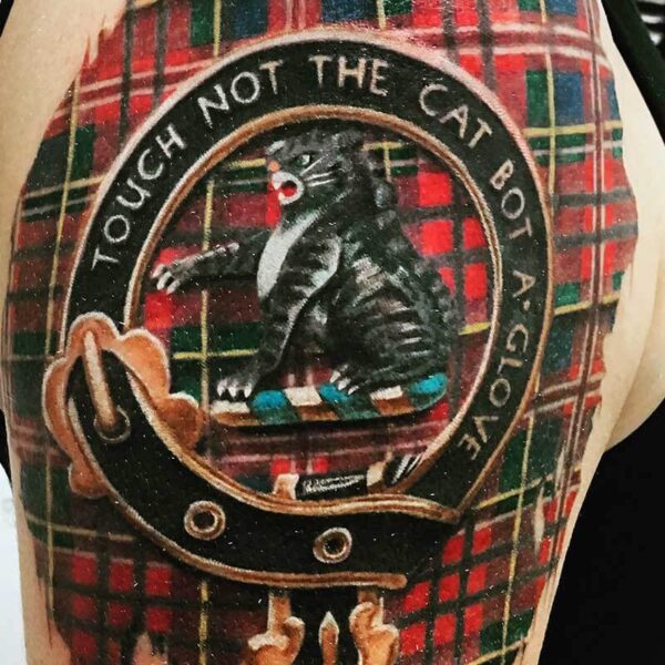Tattoo on upper arm: a circular emblem with a rat in a leather jacket inside, set against a red-green plaid background, with text around the edge.