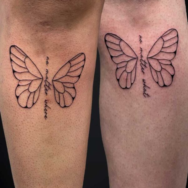 fineline-script-tattoo-butterfly-wings Two forearms display matching black butterfly-wing tattoos with vertical cursive text along the center of each winged design.