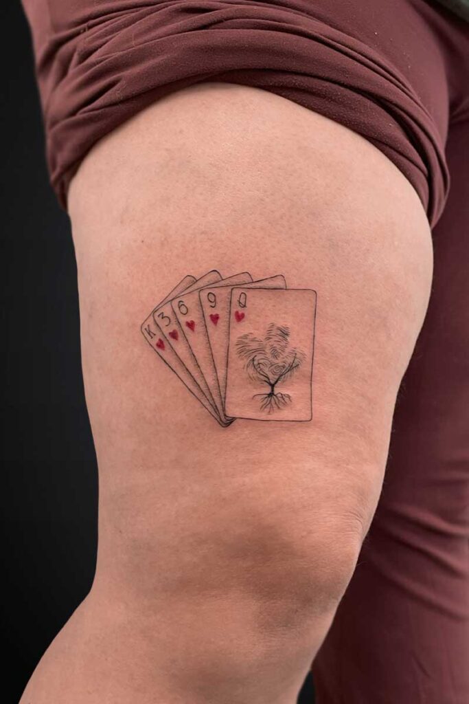 Tattoo on the thigh: five playing cards fanned out with a tree-and-root design on the rightmost card, hearts on some cards