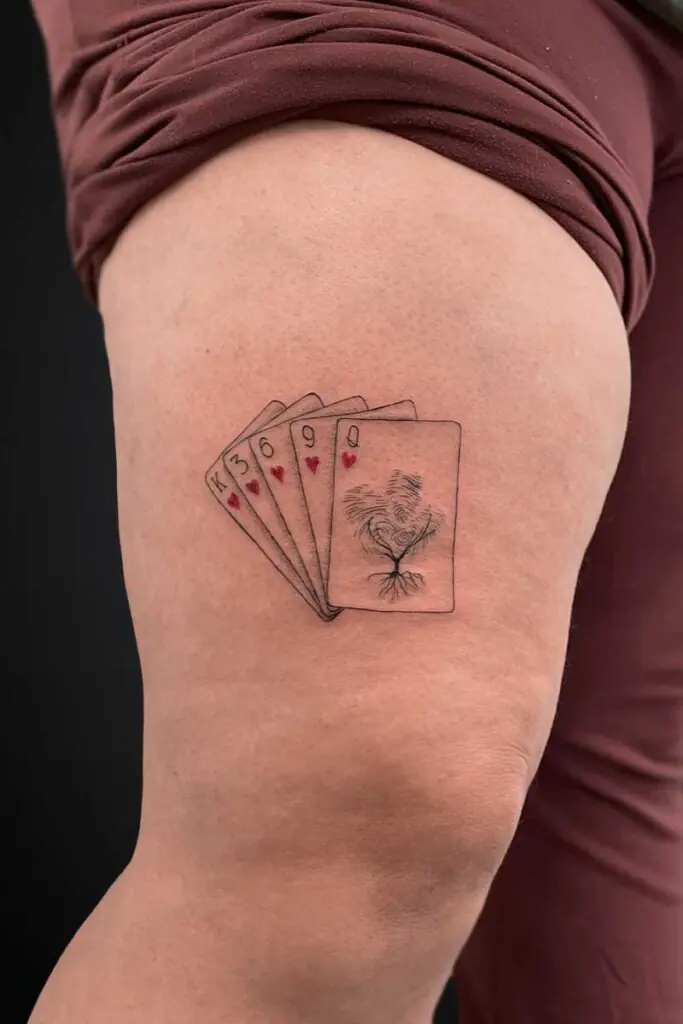 Tattoo on the thigh: five playing cards fanned out with a tree-and-root design on the rightmost card, hearts on some cards