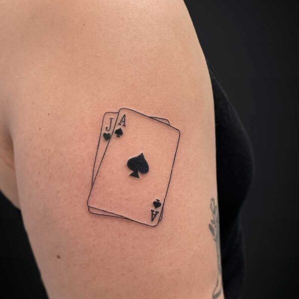 fineline-tattoo-cards-spades Tattoo on upper arm showing two overlapping playing cards: Ace of spades in front with a visible spade symbol, and Jack of spades behind it.