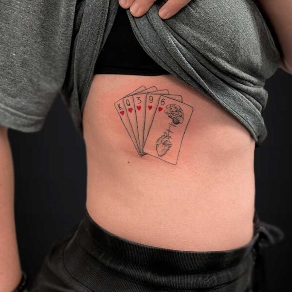 fineline-tattoo-hearts-cards Tattoo of five playing cards fanned across the person's side torso, with heart suit icons and a brain illustration on one card.