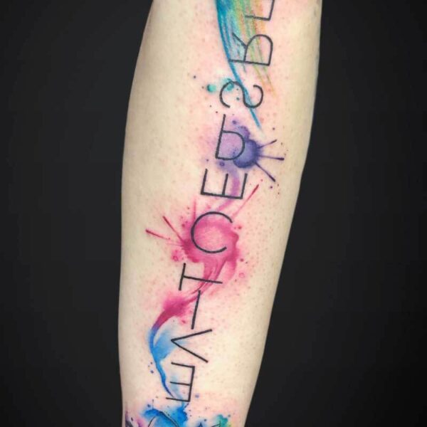 Colorful watercolor forearm tattoo with a vertical black text that reads 'LIFE BECOMES' and a lotus blossom near the wrist.
