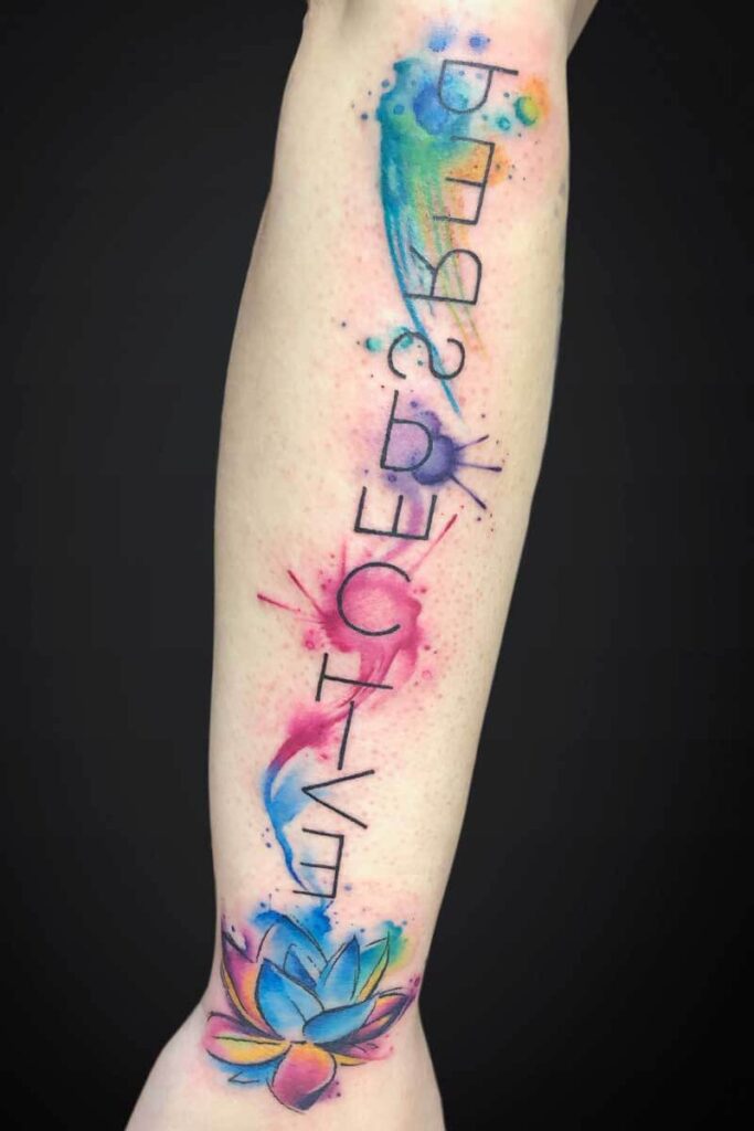 Colorful watercolor forearm tattoo with a vertical black text that reads 'LIFE BECOMES' and a lotus blossom near the wrist.