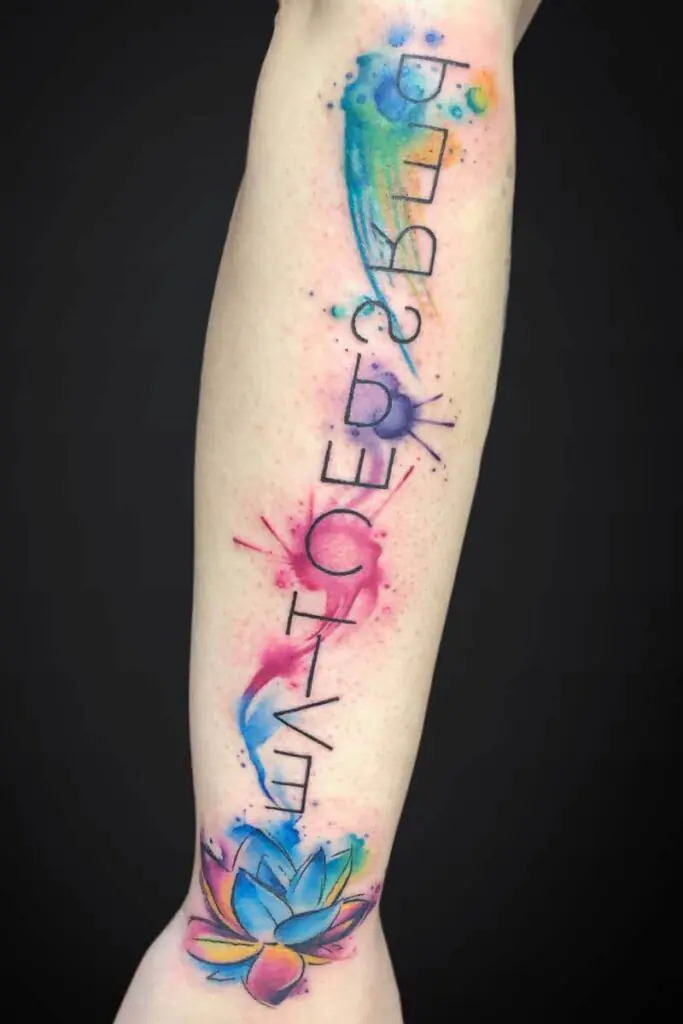 Colorful watercolor forearm tattoo with a vertical black text that reads 'LIFE BECOMES' and a lotus blossom near the wrist.