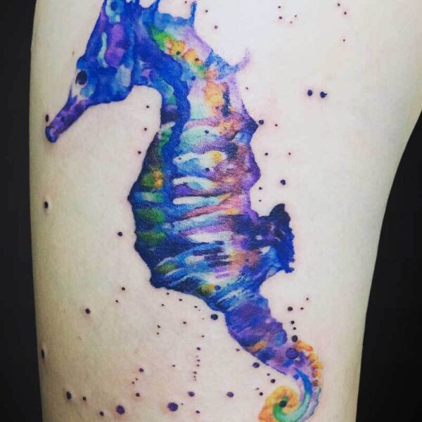 Watercolor seahorse tattoo in blue, purple, green and orange on skin, with splatter dots around it.