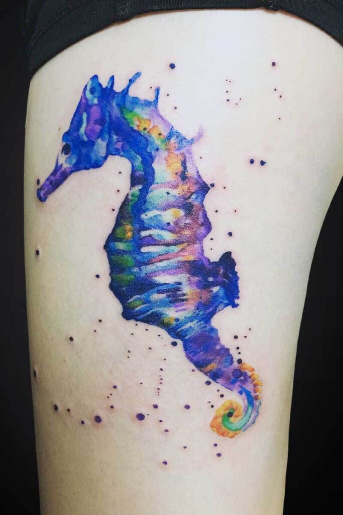 Watercolor seahorse tattoo in blue, purple, green and orange on skin, with splatter dots around it.
