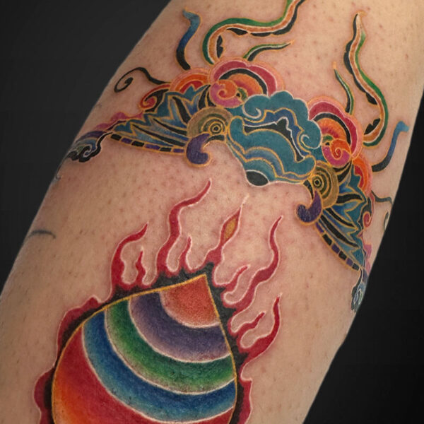 Colorful rainbow orb with red flames surrounding it, tattooed on a forearm, with decorative blue and green cloud motifs above.