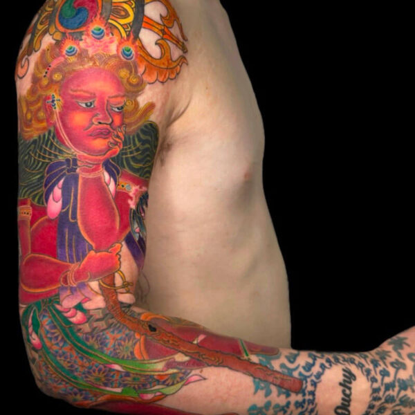 Colorful tattoo sleeve on a man’s upper arm showing a red-faced deity with an ornate headdress and flowing patterns against a black background.