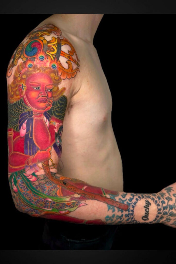 Colorful tattoo sleeve on a man’s upper arm showing a red-faced deity with an ornate headdress and flowing patterns against a black background.