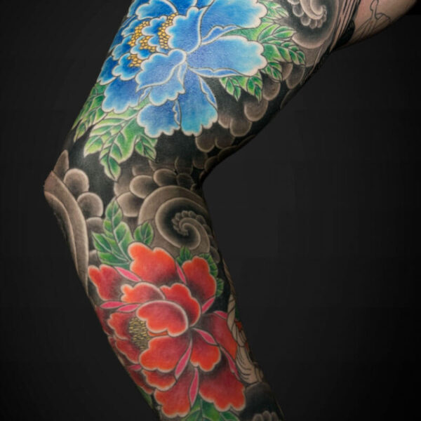 Full arm sleeve tattoo with large blue and red flowers, green leaves, and intricate gray shading along the skin.