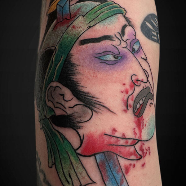 Colorful tattoo of a stylized Native American chief in profile with a green headdress, purple eye makeup, and red blood splatter on the face.