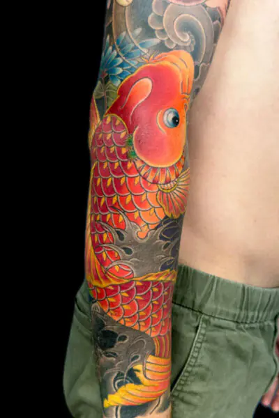 Colorful koi fish tattoo wrapping around the forearm with orange and red scales and blue accents against a dark background.
