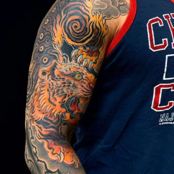 Close-up of a colorful tiger tattoo on a person's upper arm, against a dark background with a blue sleeveless shirt edge visible.