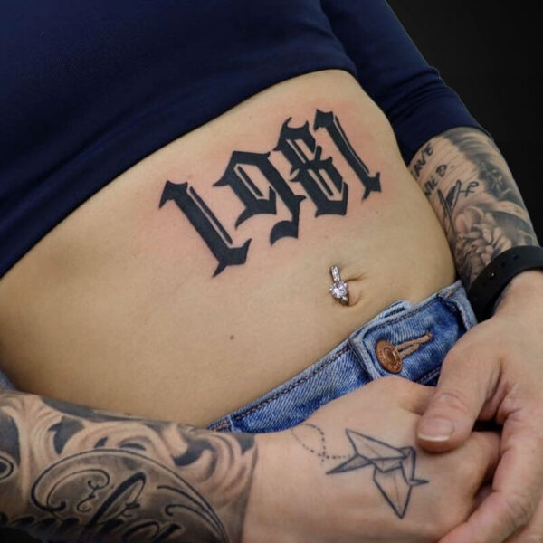 Close-up of a midriff with a large black Gothic 'LOVE' tattoo across the abdomen, plus a belly button piercing and tattooed arms.