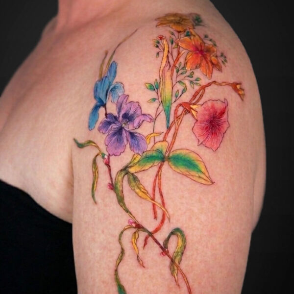 Watercolor-style tattoo of a bright, multi-colored flower bouquet running along the upper arm and shoulder, featuring blue, purple, pink, orange petals and green leaves.