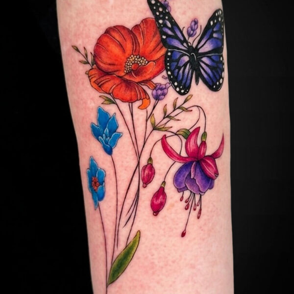 Colorful floral tattoo on a forearm with a red poppy, blue flowers, pink fuchsia, green leaves, and a blue butterfly perched on the blossoms.