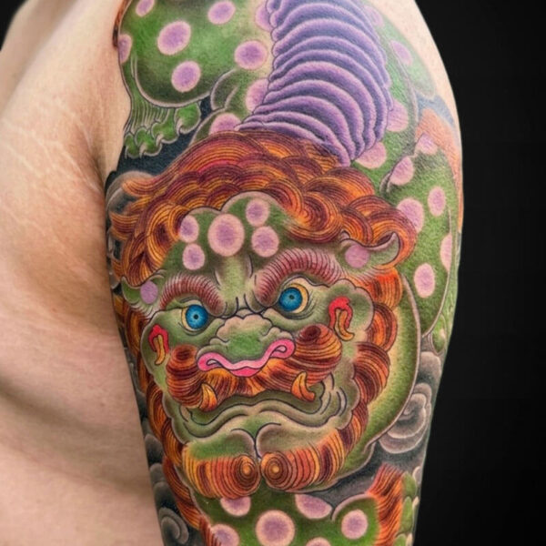 Close-up of a colorful tattoo on the upper arm: a dragon-like creature with an orange mane, green body, blue eyes, and a purple tail among gray clouds and pink spots.