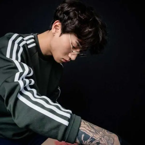 Portrait of a young man with glasses and dark hair, wearing a green striped jacket, looking down at his tattooed forearm against a dark background.