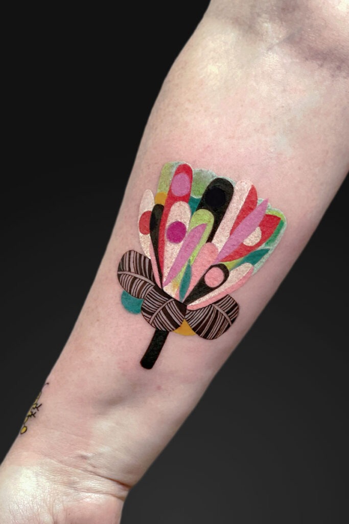Colorful abstract flower tattoo on forearm with a brown striped bow around the stem.