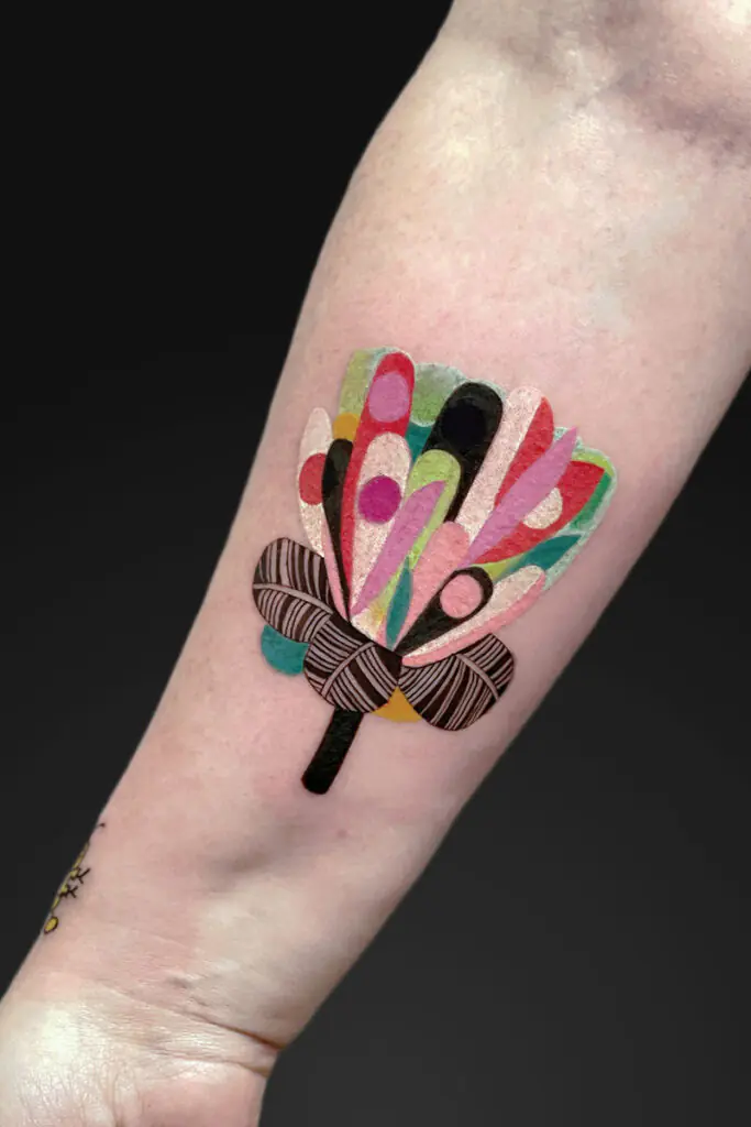Colorful abstract flower tattoo on forearm with a brown striped bow around the stem.