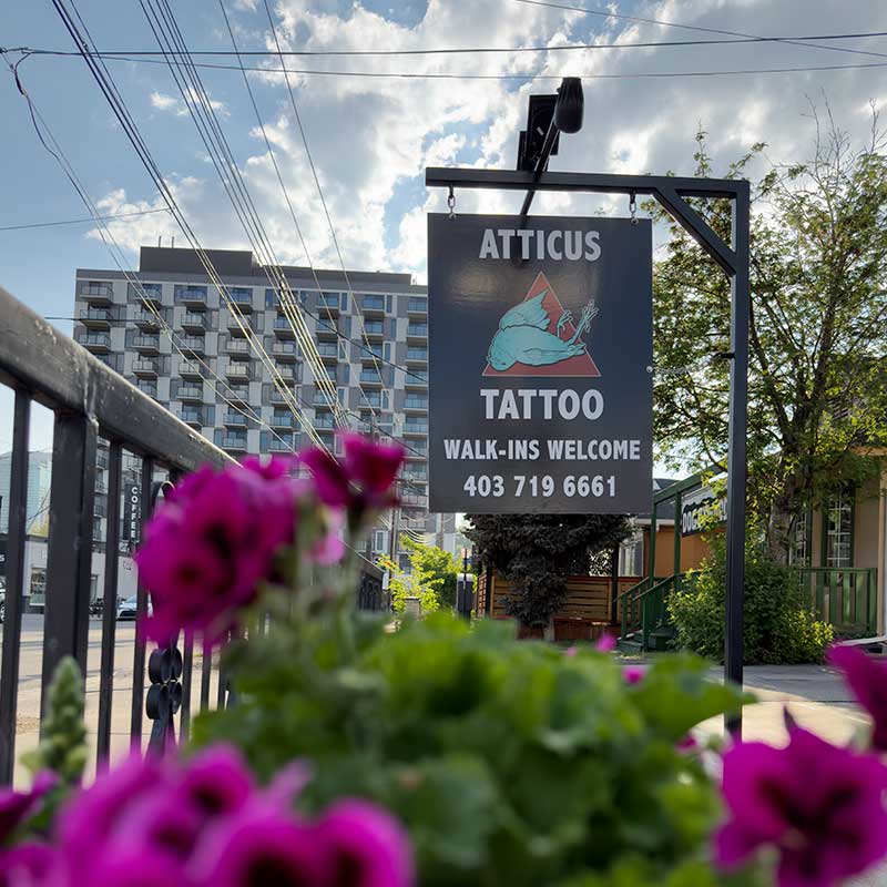 Sign for Atticus Tattoo with a blue logo on a red triangle, ‘Walk-ins Welcome’ and a phone number; urban street scene with pink flowers blur in foreground.