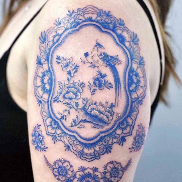 Blue tattoo on upper arm: intricate floral frame enclosing a bird among blossoms, with a decorative floral band below.