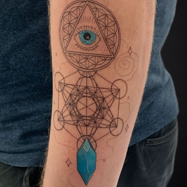 Geometric forearm tattoo with an eye inside a triangle within a circle, connected by Metatron's Cube lines and a blue crystal pendant at the bottom.