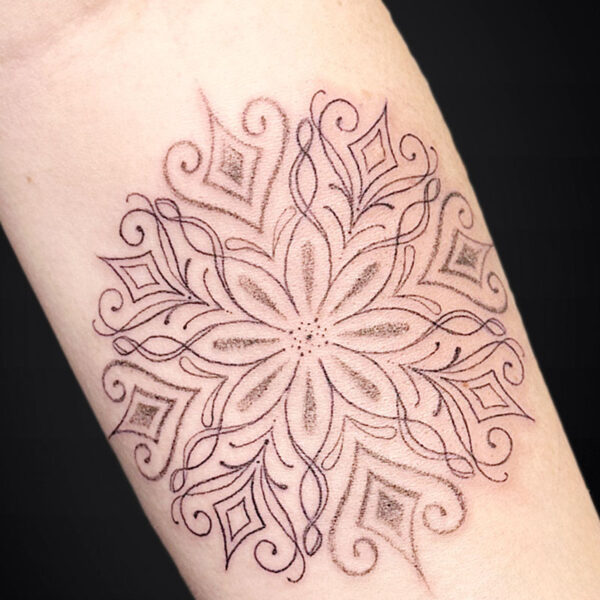 Intricate mandala-style tattoo on the forearm with petal shapes and diamond accents around a central flower motif.