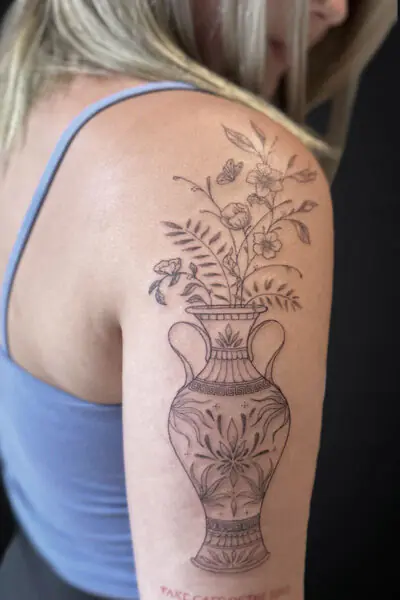 Line drawing tattoo of an ornate vase with blooming flowers on a person's upper arm.