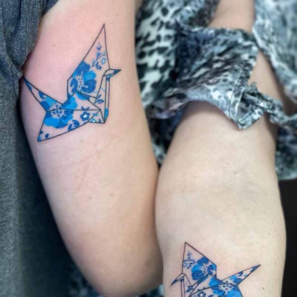 Two forearms showing matching origami crane tattoos with blue floral patterns on pale skin.