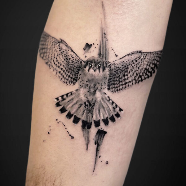 Black and gray tattoo of a bird of prey in flight across the forearm, wings spread wide and tail fanned, with watercolor splashes and geometric line accents around the body.