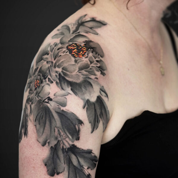 Grayscale floral tattoo on a woman's upper arm and shoulder, featuring large peony blossoms with small orange centers.
