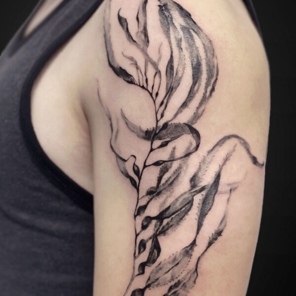 Black and gray botanical tattoo along the upper arm, resembling a flowing, leaf-like wing with shaded petals and curved lines.