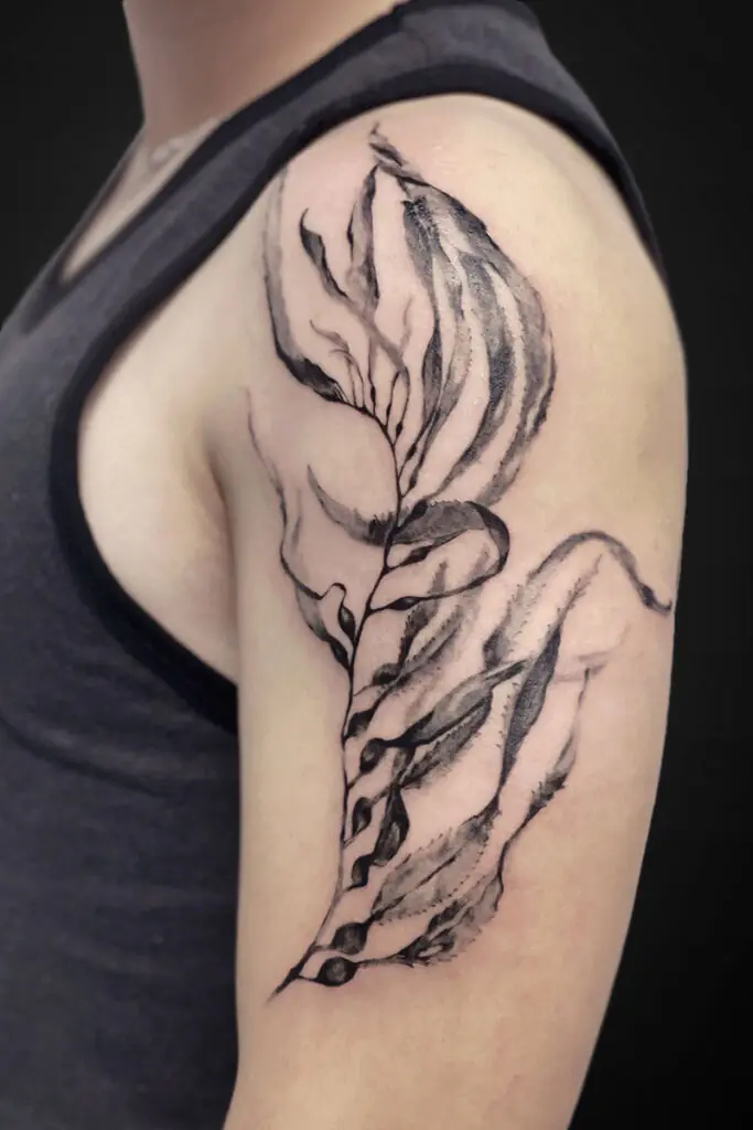 Black and gray botanical tattoo along the upper arm, resembling a flowing, leaf-like wing with shaded petals and curved lines.