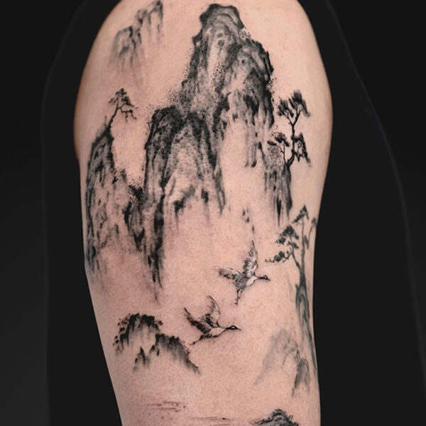 Sumi-e style tattoo on the upper arm: jagged mountains, mist, a calm lake, and birds in flight.