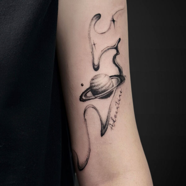 Black ink forearm tattoo of Saturn with a ring and decorative swirling ribbons, plus the cursive word 'Adventure'.