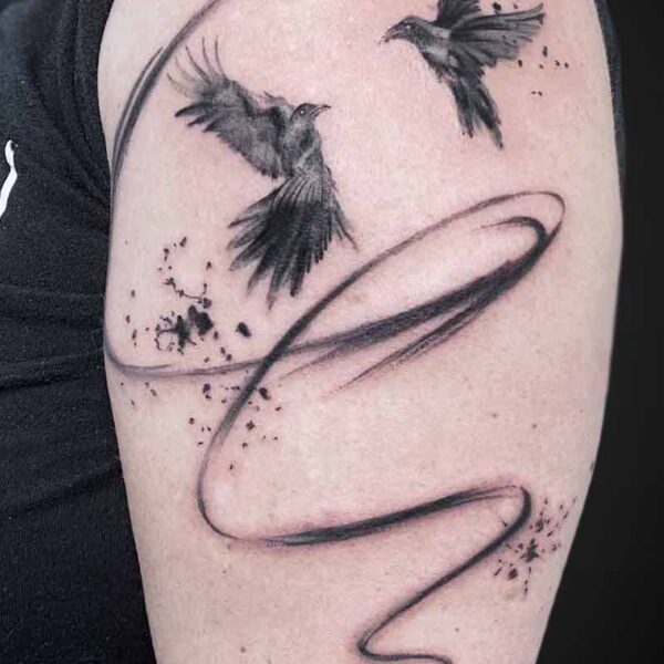 Tattoo of two small birds in flight above a sweeping curved line with ink splatters on the upper arm.
