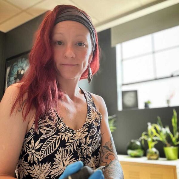 Woman with red hair wearing a black headband and a tropical leaf-pattern tank top, standing indoors with potted plants in the background, slight smile.