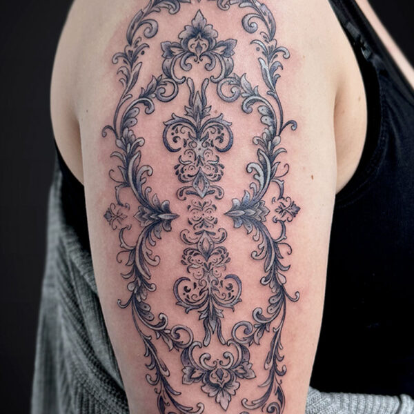 ornamental-tattoo-floral-decorative-elements Intricate black-and-gray ornamental tattoo running from shoulder to upper arm, featuring symmetrical scrollwork and floral motifs.