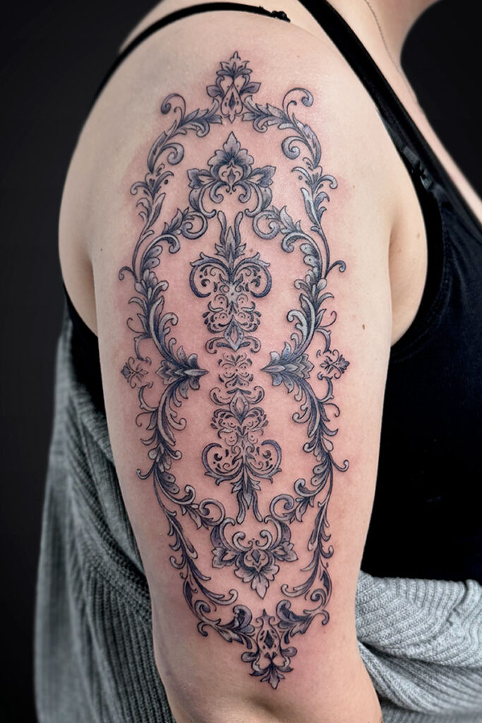 Intricate black-and-gray ornamental tattoo running from shoulder to upper arm, featuring symmetrical scrollwork and floral motifs.