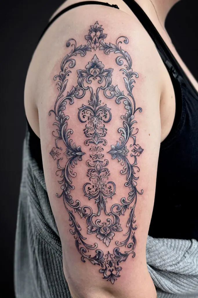 Intricate black-and-gray ornamental tattoo running from shoulder to upper arm, featuring symmetrical scrollwork and floral motifs.