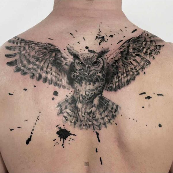 Back tattoo of a detailed owl with wings spread wide, black ink, with splash-style accents across the shoulders and upper back.