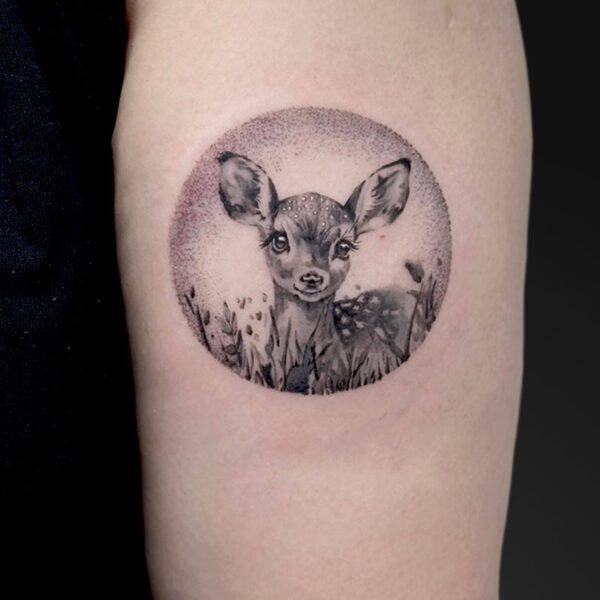 Circular grayscale tattoo of a fawn's head peeking through grass, inside a dotted circle frame in soft shading.