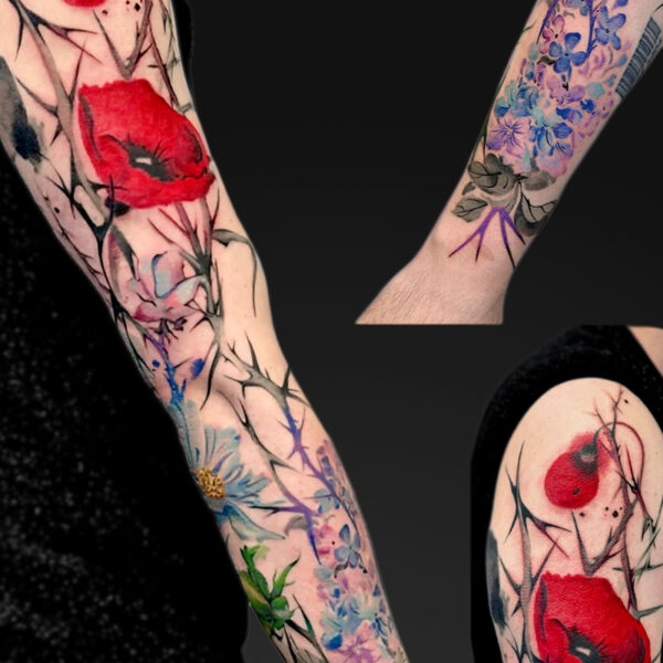 Colorful floral tattoo sleeves on forearms, featuring red poppies, blue blossoms, and black branch details on light skin against a dark background.