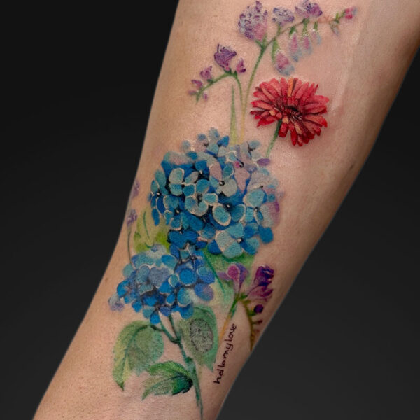 Watercolor-style forearm tattoo of blue hydrangea clusters with purple blossoms and a red bloom, green stems, and small script text along the stem.