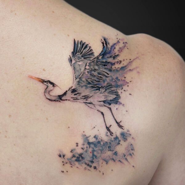 Watercolor-style tattoo of a heron in flight on a person’s upper back shoulder, with blue-purple splashes behind.