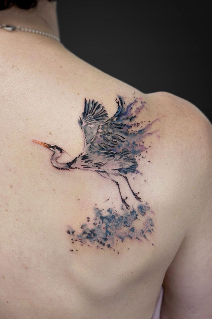 Watercolor-style tattoo of a heron in flight on a person’s upper back shoulder, with blue-purple splashes behind.